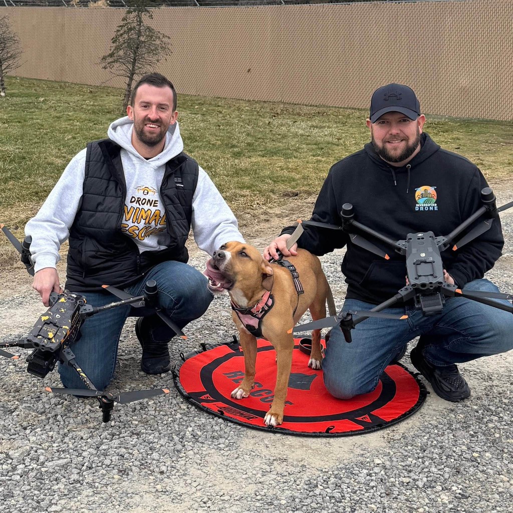 Two drone pilots kneeling with a dog and their drones.